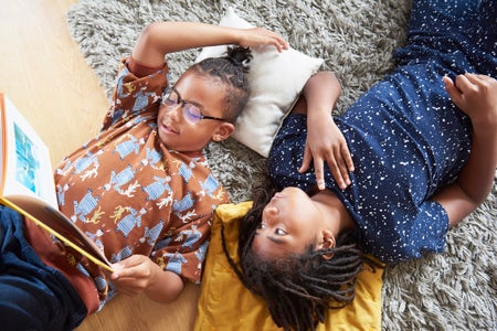 A child reads a book while lying on a rug, while another child looks on.