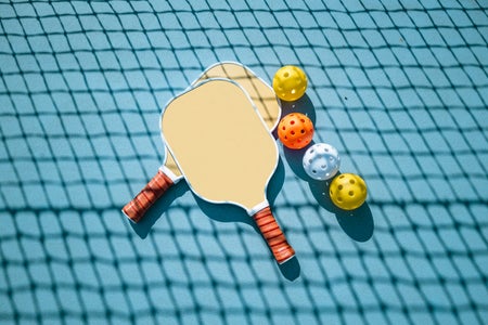 A still life showing pickleball paddles, balls and net on a colorful light blue background.