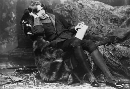 A man in Victorian clothing lounges on a fur-draped couch, resting his head on his right hand.