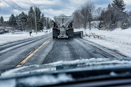 Car driver perspective looking through windshield while following a large plow truck spreading rock salt on a rural highway during a snow storm.