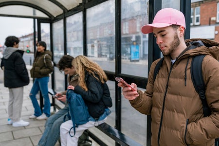 A shot of teenage commuters standing and waiting for the bus .There are two groups of friends and one man standing by himself using his phone.