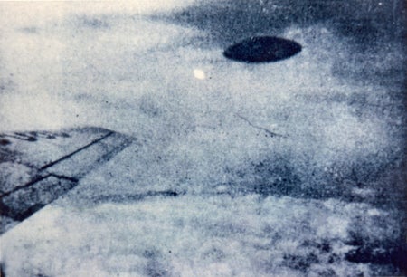 A black disk near the wingtip of an airplane, with clouds below.