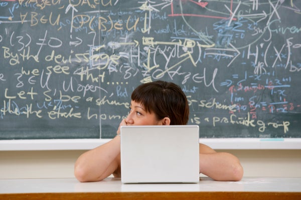 A person sits at a desk with their face partially obscured by a laptop in front of a chalkboard covered in mathematical equations