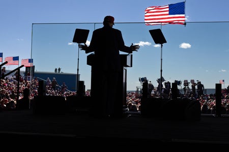 Republican presidential nominee  and former president Donald Trump, photographed from behind in silhouette, speaks from behind a glass barrier. In the distance beyond the silhouette of Trump, podium and stage, the audience and several American flags can be seen