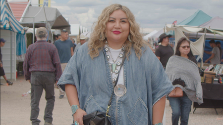 A woman faces the viewer smiling slightly, dressed in a blue to and jewelry walking through a crowd